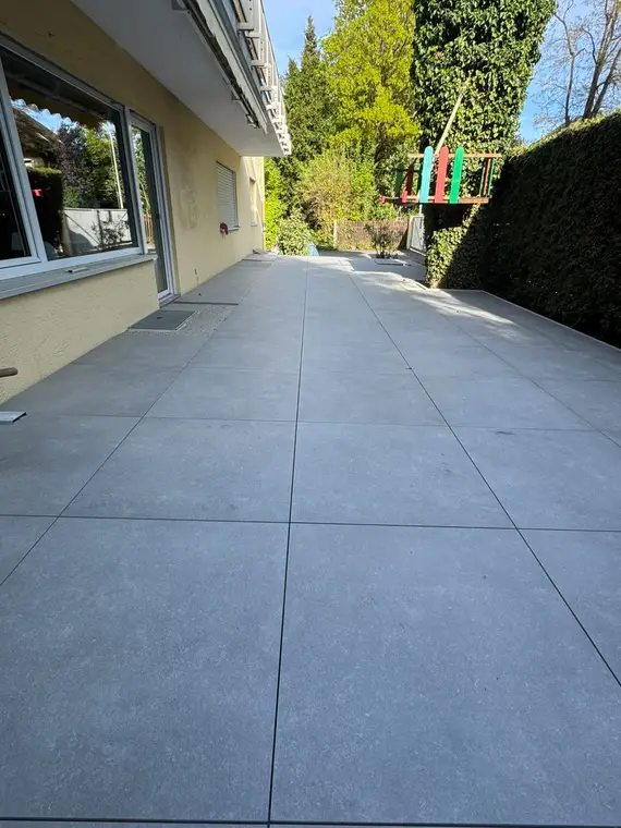 Neugepflasterte Terrasse mit grauen Fliesen neben Wohnhaus und Garten