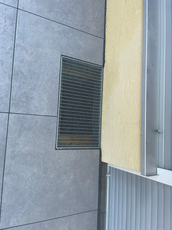 Gitterrost im Fliesenboden neben einer gelben Hauswand mit Lichtschacht